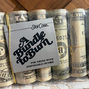 May include: A bundle of five cylindrical rolls wrapped in clear plastic, each featuring the print of United States dollar bills. A square gray tag is attached with gold string, reading StarCase A bundle to Burn For those with disposable income.