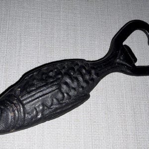 May include: A black, cast iron bottle opener shaped like a fish. The body of the fish features a textured, scale-like pattern across the surface, while the tail end of the fish forms the functional loop and hook mechanism for opening bottles.