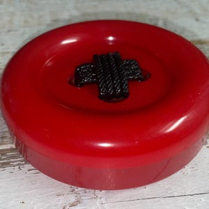 May include: A large, round, red button with a black cross-shaped design in the center. The button has a glossy finish and a raised edge. The button is a sewing or craft supply.