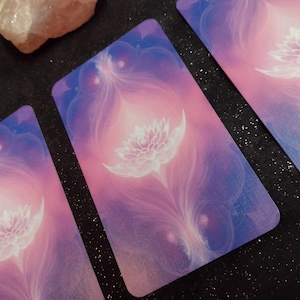 May include: Three tarot cards with a white lotus flower design on a purple and pink background.