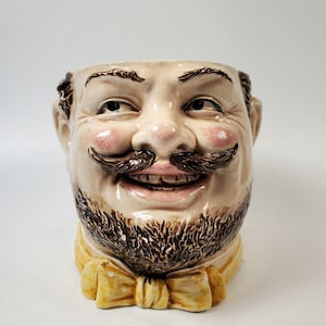 Puede incluir: Taza de cerámica con forma de rostro de hombre sonriente. La taza presenta un rostro detallado con bigote, barba y pajarita amarilla. La cara tiene mejillas rosadas y cabello castaño.