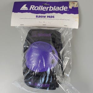 Puede incluir: Coderas Rollerblade en un embalaje de plástico transparente. Las almohadillas tienen una carcasa dura púrpura y un respaldo de tela negra. El embalaje incluye el logotipo de Rollerblade y el texto "ELBOW PADS".
