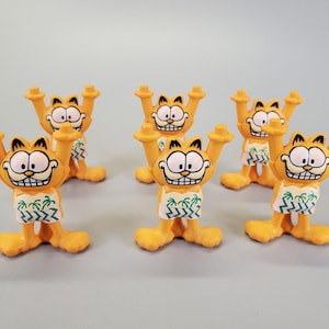 May include: Six orange Garfield figurines with arms raised, wearing white and green patterned shorts. The figurines have black and white facial features and are standing on a light blue surface.