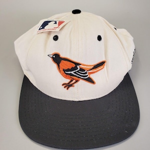May include: A baseball cap with a cream-coloured crown and a dark grey brim. The cap features an embroidered orange and black bird logo and a tag with the MLB logo. The cap has a classic, structured design.