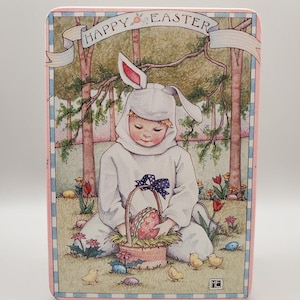 May include: A rectangular Easter-themed box with a pink border and a decorative illustration. The illustration depicts a child in a bunny costume holding a basket with an Easter egg. The words "HAPPY EASTER" are written across the top.