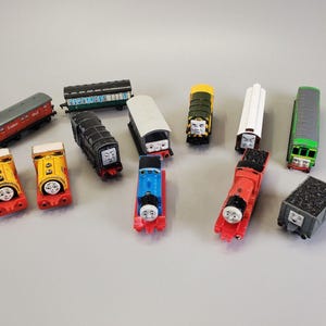 Op de afbeelding: Een verzameling kleurrijke speelgoedtreinlocomotieven en -wagons, waaronder rode, gele, blauwe, groene en zwarte modellen. Elke trein heeft een lachend gezicht en de set bevat een kolenwagen en passagierswagons.