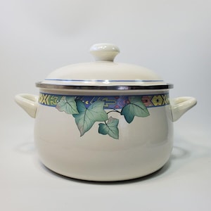 Op de afbeelding: Crème-kleurige emaille kookpot met deksel. De pot heeft een decoratieve band met geometrische patronen en groene bladeren. Het heeft twee handvatten aan de zijkant en een witte knop op het deksel. Geschikt om te koken.