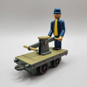 May include: A classic toy train flatbed carriage with a figure of a man in a blue suit, yellow tie, and bowler hat. The flatbed is olive green with dark brown wheels. The man is standing near a mechanical device.