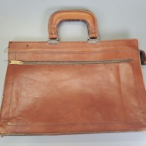 May include: A vintage brown leather briefcase with a top handle and a gold-coloured zip closure. The briefcase has a rectangular shape and shows signs of wear, adding to its classic appeal. The handle is also made of brown leather.