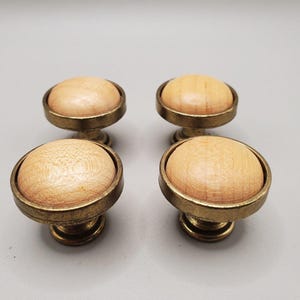Peut inclure: Quatre boutons de placard circulaires disposés en carré. Chaque bouton présente un insert central en bois naturel clair et lisse, enchâssé dans un cadre circulaire en métal de couleur laiton foncé avec une base étagée.