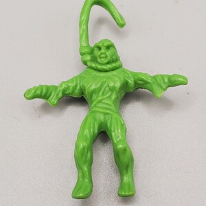 May include: A bright green plastic figurine of a humanoid creature with a textured, muscular body and a wide, open-mouthed expression. The figure features a prominent, curved hook-like appendage extending upward from the neck area, resembling a rope.