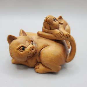 May include: A light brown wooden carving of a cat lying down with a small mouse resting on its back. The cat has pointed ears and dark eyes, while the mouse is positioned near the cat's tail, which curves upward along the mouse's body.