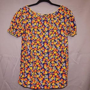 May include: A short-sleeved top with a colourful floral design. The navy blue material is patterned with small yellow, orange, and white flowers. The neckline and sleeve edges have a yellow trim. The top is suspended from a black hanger.