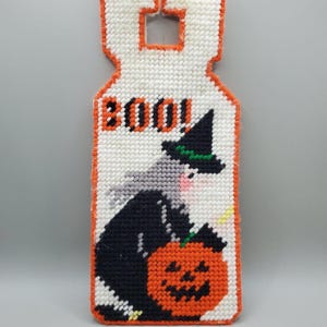 May include: A plastic canvas door hanger featuring a stitched design of a witch with gray hair and a black hat holding an orange jack-o'-lantern. The word BOO! is stitched in black at the top. The entire piece is edged in bright orange yarn.