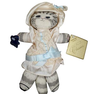 Puede incluir: Una muñeca de gato de peluche que lleva un vestido color crema con encaje y un lazo azul claro. La muñeca tiene un sombrero a juego y piernas a rayas grises y blancas. Una etiqueta dice "Classics."