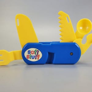 Peut inclure: Un outil multifonction en plastique bleu avec des accessoires jaunes comprenant une fourchette, une lame dentelée, une cuillère et un décapsuleur. Le manche bleu porte un autocollant ovale blanc avec le texte coloré soft stuff.