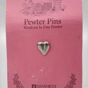 Puede incluir: Un alfiler de peltre en forma de corazón sobre una tarjeta rosa. La tarjeta dice "Pewter Pins" y "Handcast In Fine Pewter". El alfiler es de Danforth Pewterers, Maiden Vermont.