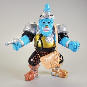May include: A blue action figure with a silver helmet and shoulder pads, a gold and black torso, and brown accents. The figure has a small tan satchel and a silver ray gun. The figure is posed with arms outstretched.