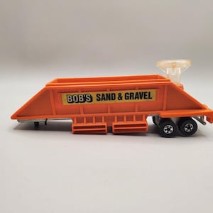 May include: An orange toy dump trailer with a yellow rectangular decal on the side featuring the black text BOB'S SAND & GRAVEL. The trailer has a clear plastic hitch component at the front and two sets of black wheels with silver centers.