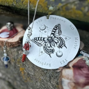 May include: A silver pendulum with a red crystal point hangs from a silver chain. The pendulum is resting on a silver circular divination board with a black and white butterfly design. The board has the words "Yes", "Maybe", "No", "Unknown", and "Rephrase" printed around the edge.