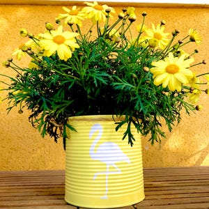 Könnte beinhalten: Ein Arrangement aus gelben Gänseblümchen und grünen Blättern in einem recycelten gelben Dosenpflanzgefäß. Die Dose ist mit einer weißen Flamingosilhouette verziert. Die Blumen blühen in voller Blüte und schaffen eine fröhliche und dekorative Darstellung.