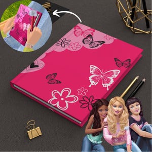 May include: Pink notebook with a butterfly design and a heart. The notebook has a pen holder and is closed. Three dolls are in the background.