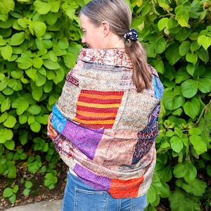 May include: A woman wearing a colourful patchwork scarf with a variety of patterns and colours, including red, orange, purple, and blue. The scarf is made of a soft, flowing fabric and is draped over the woman's shoulders.