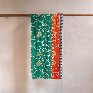 May include: A green and orange kantha quilt with a floral pattern hangs on a wooden rod. The quilt has a white border and a blue trim.