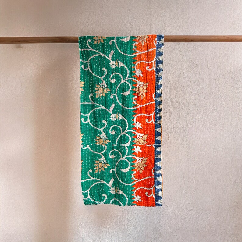 May include: A green and orange kantha quilt with a floral pattern hangs on a wooden rod. The quilt has a white border and a blue trim.