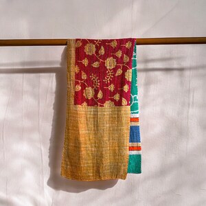 May include: A multi-colored kantha quilt with a floral pattern in shades of red, gold, and green. The quilt is hanging on a wooden rod.
