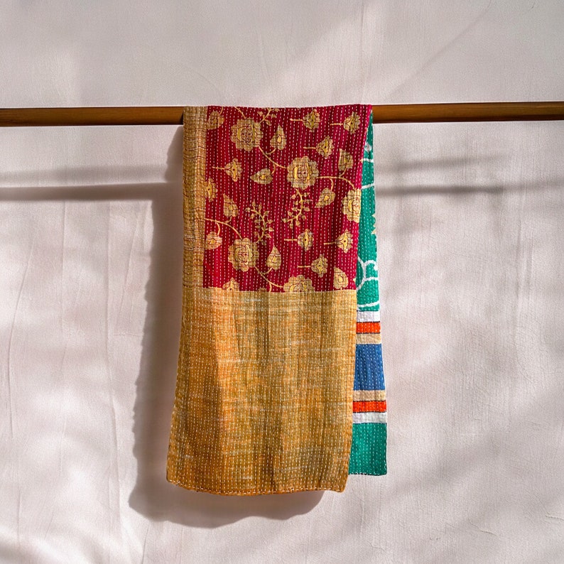 May include: A multi-colored kantha quilt with a floral pattern in shades of red, gold, and green. The quilt is hanging on a wooden rod.