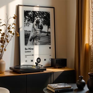 Könnte beinhalten: Ein gerahmter Schwarz-Weiß-Foto-Druck mit dem Text "Be Mine" und einer Musikplayer-Oberfläche. Ein Plattenspieler steht auf einem Holzschrank, mit einer Vase mit getrockneten Zweigen und anderen dekorativen Gegenständen.