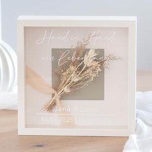 Könnte beinhalten: Ein weißer Schattenrahmen mit einem Strauß getrockneter Blumen und dem Text "Hand in Hand ein Leben lang". Die Namen "Jana & Leon" und zwei Daten sind ebenfalls sichtbar. Der Strauß ist mit einem beigen Band gebunden.