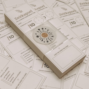 May include: A stack of white paper cards with German text, including "Zeitkapsel" and "10". A clear plastic band wraps the stack, with a circular sticker featuring a sun design. The cards are for recording memories.