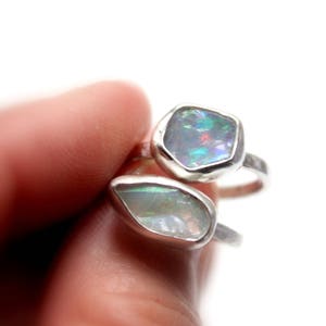 May include: Two silver rings with opal gemstones. The top ring features a hexagonal opal with a rainbow of colors. The bottom ring features a teardrop-shaped opal with a milky white color.