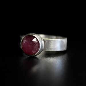 May include: A silver ring with a round, faceted, pink ruby gemstone set in a bezel setting.