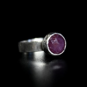 May include: A silver ring with a round, faceted, pink ruby gemstone set in a bezel setting.