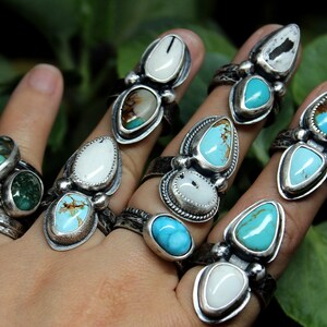 May include: A hand wearing multiple silver rings with turquoise and white stones. The rings are stacked on each finger and feature different designs.