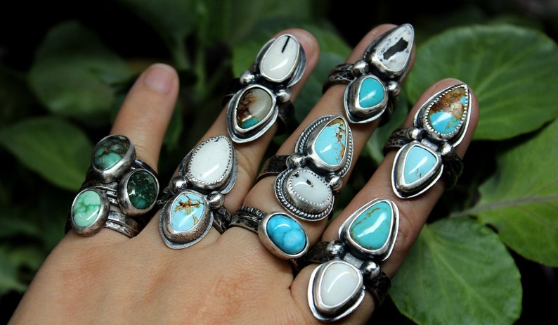 May include: A hand wearing multiple silver rings with turquoise and white stones. The rings are stacked on each finger and feature different designs.