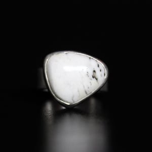 Può includere: Un anello d'argento con una pietra bianca triangolare. La pietra presenta macchie nere ed è incastonata in una lunetta d'argento. L'anello è presentato su sfondo nero, evidenziando il design del gioiello.