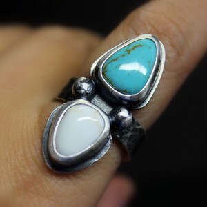 May include: A silver ring with two teardrop-shaped stones. The top stone is turquoise blue, and the bottom stone is white.