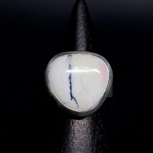Può includere: Un anello d'argento con una grande pietra di forma ovale. La pietra è principalmente bianca con venature rosa e nere. L'anello è posto su uno sfondo nero, evidenziando il suo design e colore.