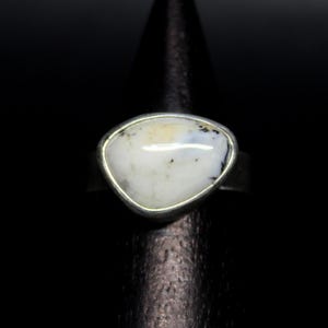 Può includere: Un anello d'argento con una grande pietra bianca di forma irregolare con inclusioni nere e gialle. La pietra è incastonata in una semplice fascia d'argento, creando un contrasto sorprendente sullo sfondo scuro. L'anello è esposto su un supporto nero.