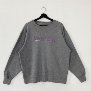 May include: A gray crew neck sweatshirt with the text "Agnes Scott College" in purple and white. The sweatshirt is hanging on a black hanger against a white background. The sleeves have ribbed cuffs.