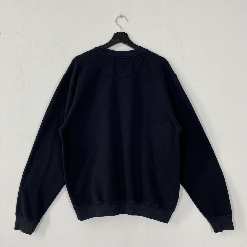 May include: A black crew neck sweatshirt hanging on a black hanger against a white wall. The sweatshirt has long sleeves and a ribbed hem and cuffs. The back of the sweatshirt is visible, showing a plain design.