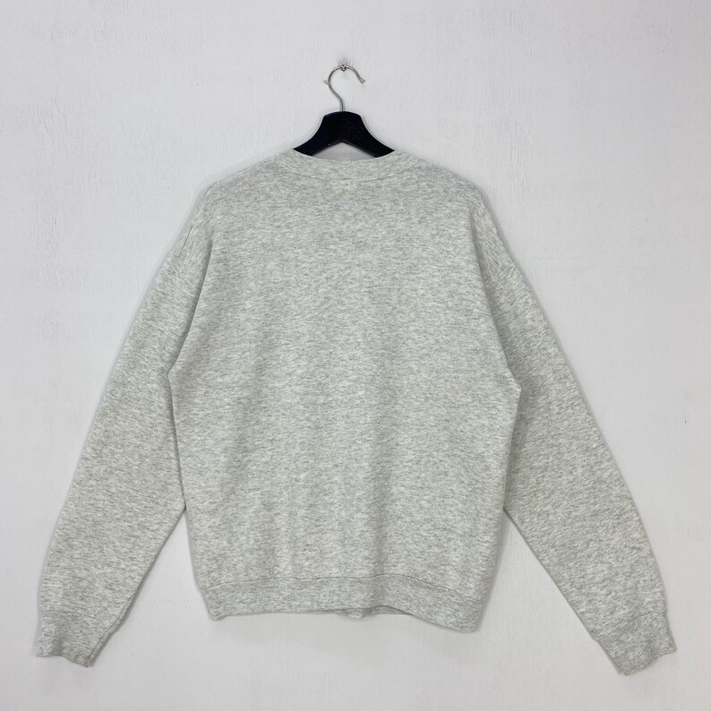 Może przedstawiać: Szara melanżowa bluza dresowa wisi na czarnym wieszaku na białym tle. Długi rękaw ma okrągły dekolt i miękką, nakrapianą fakturę. Bluza to codzienna, swobodna odzież.