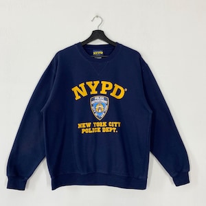 Peut inclure: Sweat-shirt bleu marine NYPD avec le logo NYPD jaune et un écusson de la police. Le texte "NEW YORK CITY POLICE DEPT." est en dessous du logo. Le sweat-shirt est suspendu à un cintre noir.