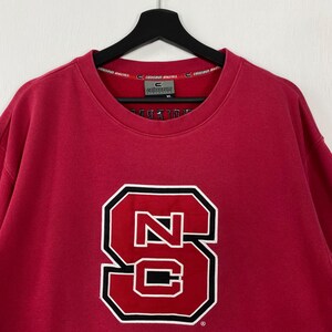 K&ouml;nnte beinhalten: Ein rotes Sweatshirt mit einem gro&szlig;en, wei&szlig; umrandeten, schwarz umrandeten "NC" Logo. Das Sweatshirt hat einen Rundhalsausschnitt und lange &Auml;rmel. Der Markenname "Colosseum Athletics" ist auf dem Kragen aufgedruckt.