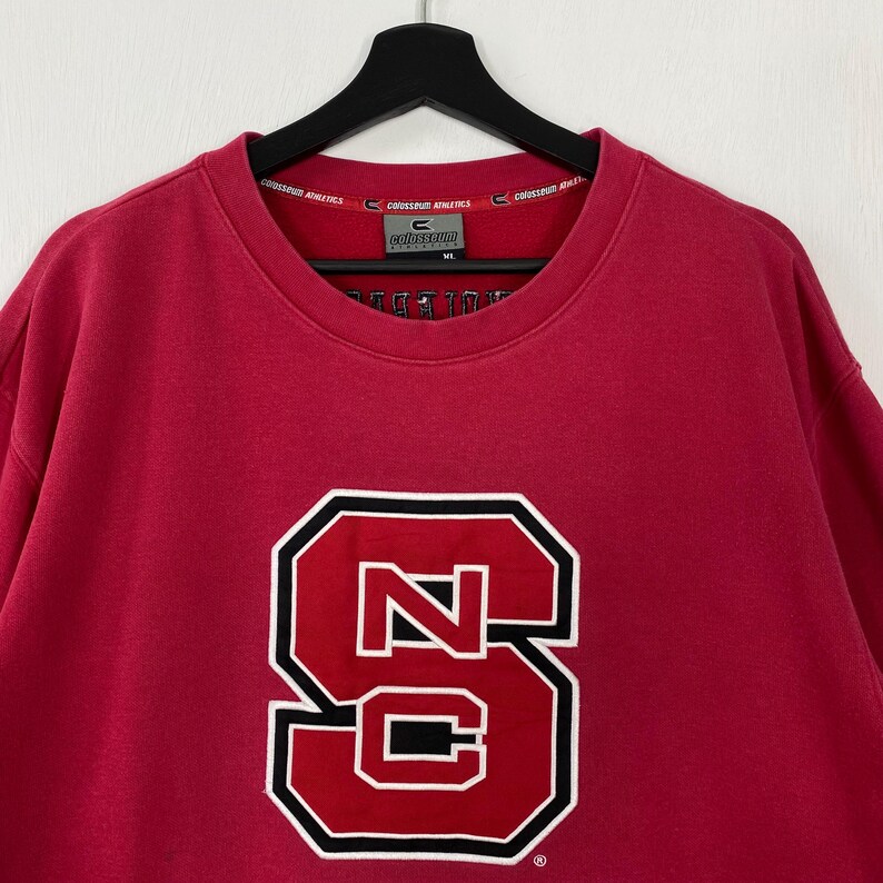 K&ouml;nnte beinhalten: Ein rotes Sweatshirt mit einem gro&szlig;en, wei&szlig; umrandeten, schwarz umrandeten "NC" Logo. Das Sweatshirt hat einen Rundhalsausschnitt und lange &Auml;rmel. Der Markenname "Colosseum Athletics" ist auf dem Kragen aufgedruckt.