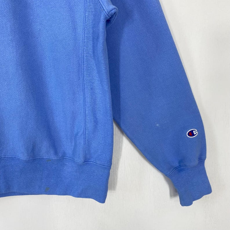 Peut inclure: Un sweat-shirt Champion bleu clair. Le sweat-shirt a un col rond et des manches longues. Le logo Champion est visible sur le poignet de la manche gauche. Le tissu semble &ecirc;tre une mati&egrave;re douce et textur&eacute;e.